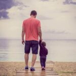 Father holding toddler daughter's hand while walking towards water.