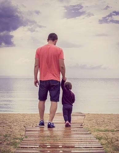 Father holding toddler daughter's hand while walking towards water.