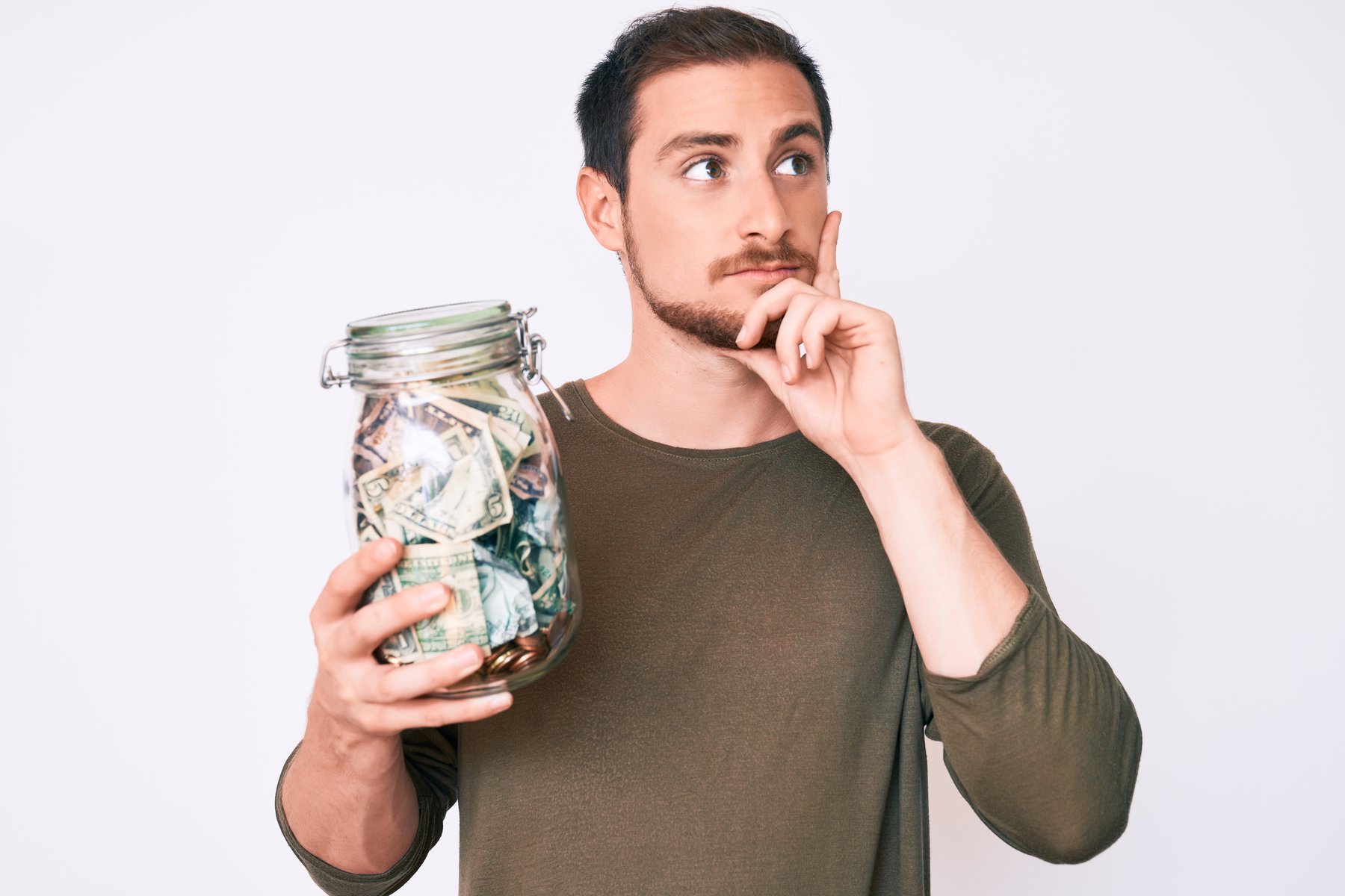 Young handsome man holding jar with savings serious face thinking about question with hand on chin, thoughtful about confusing idea